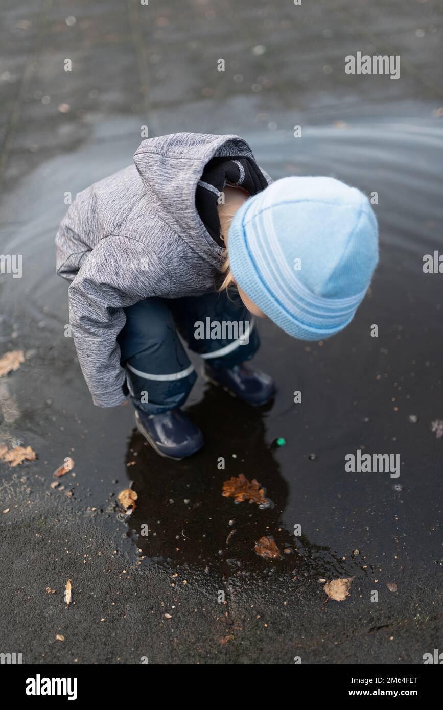 Blick von oben auf einen zweijährigen Jungen in Regenhosen und Gummistiefeln, der nach einer Regendusche in der Wasserpfütze spielt Stockfoto