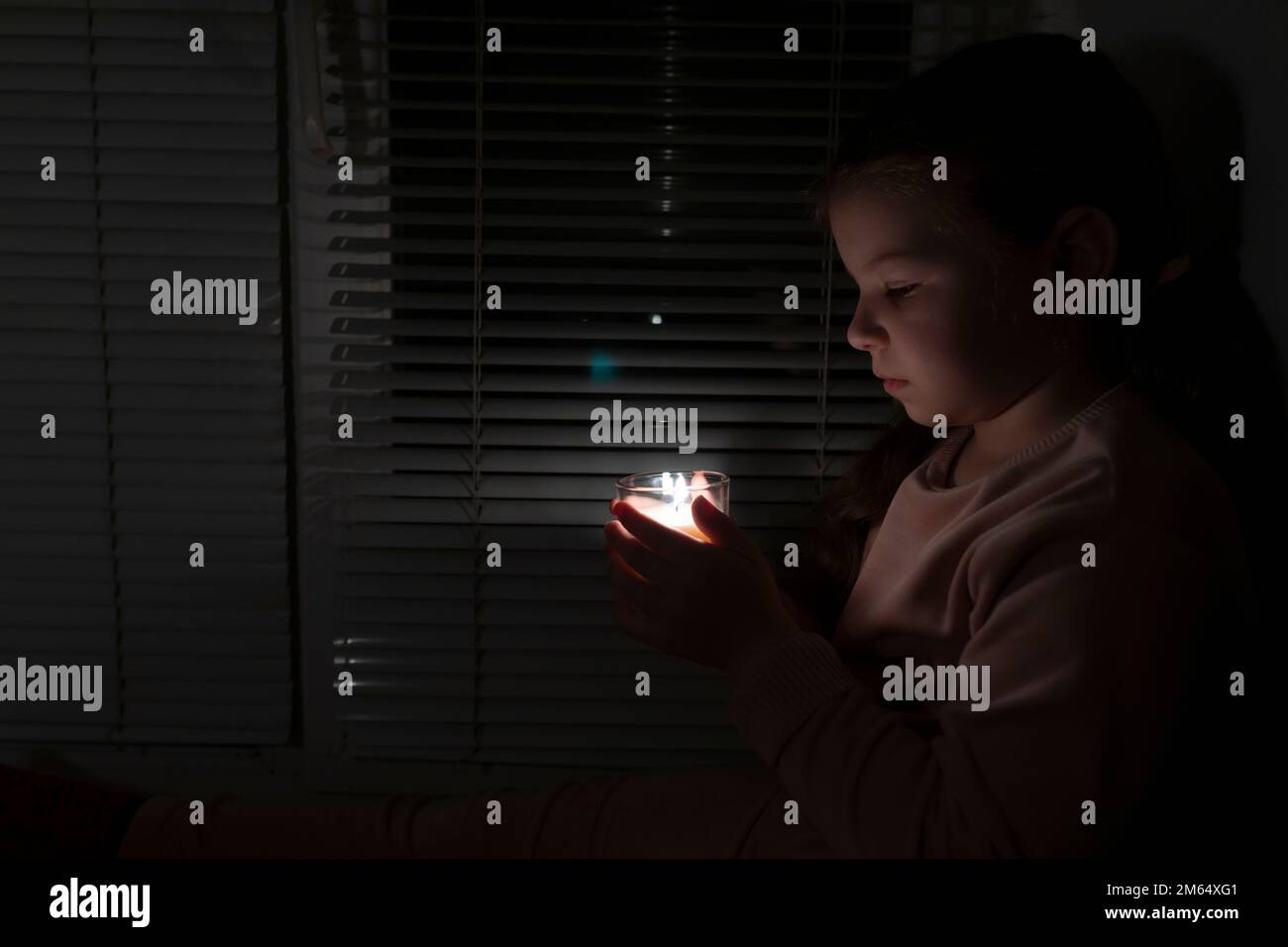 Ein Mädchen in einem dunklen Raum nahe dem Fenster hält eine Kerze in einem Glas in der Hand. Stromausfallkonzept. Stromausfall. Energiekrise Stockfoto Ein Mädchen in einem dunklen Raum nahe dem Fenster hält eine Kerze in einem Glas in der Hand. Stromausfallkonzept. Stromausfall. Energiekrise Stockfoto