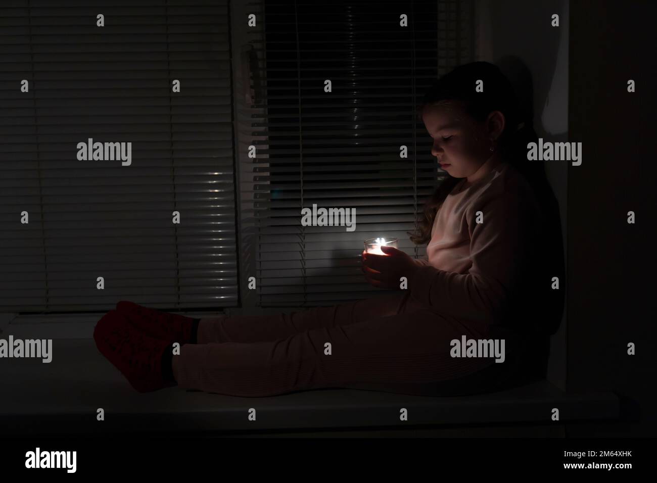 Ein Mädchen in einem dunklen Raum nahe dem Fenster hält eine Kerze in einem Glas in der Hand. Stromausfallkonzept. Stromausfall. Energiekrise Stockfoto Ein Mädchen in einem dunklen Raum nahe dem Fenster hält eine Kerze in einem Glas in der Hand. Stromausfallkonzept. Stromausfall. Energiekrise Stockfoto