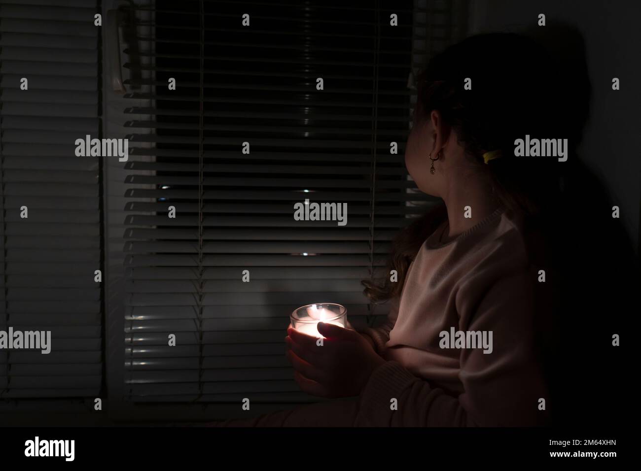 Ein Mädchen in einem dunklen Raum nahe dem Fenster hält eine Kerze in einem Glas in der Hand. Stromausfallkonzept. Stromausfall. Energiekrise Stockfoto Ein Mädchen in einem dunklen Raum nahe dem Fenster hält eine Kerze in einem Glas in der Hand. Stromausfallkonzept. Stromausfall. Energiekrise Stockfoto