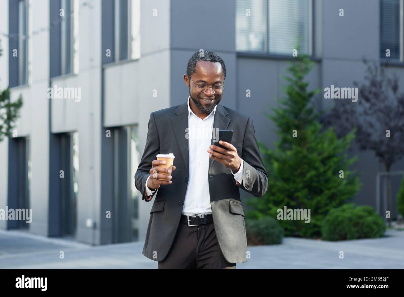 Ein erwachsener Geschäftsmann in einem Business-Anzug, der sein Telefon außerhalb des Bürogebäudes benutzt, ein afroamerikanischer Chef hält einen Becher heißes Getränk, lächelt, schreibt SMS und surft auf Websites. Stockfoto