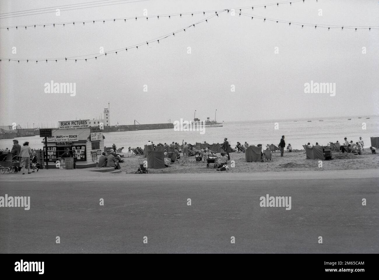 1950er, historisch, Menschen am Strand von North Bay in Scarborough, einem Strandresort in East Yorkshire, England, Großbritannien. Ein Kiosk dort "Beach Pictures" bietet Happy Snaps on the Beach, produziert von Photo Casino Ltd von Scarborough & Clayton Bay. Die Küstenstadt entwickelte sich zu einem wichtigen Reiseziel am Meer, und viele Fotostudios wurden eingerichtet, um den Besuchern Urlaubsfotos zu liefern, einschließlich nach dem Zweiten Weltkrieg, Photo Casino Co. Ltd Stockfoto