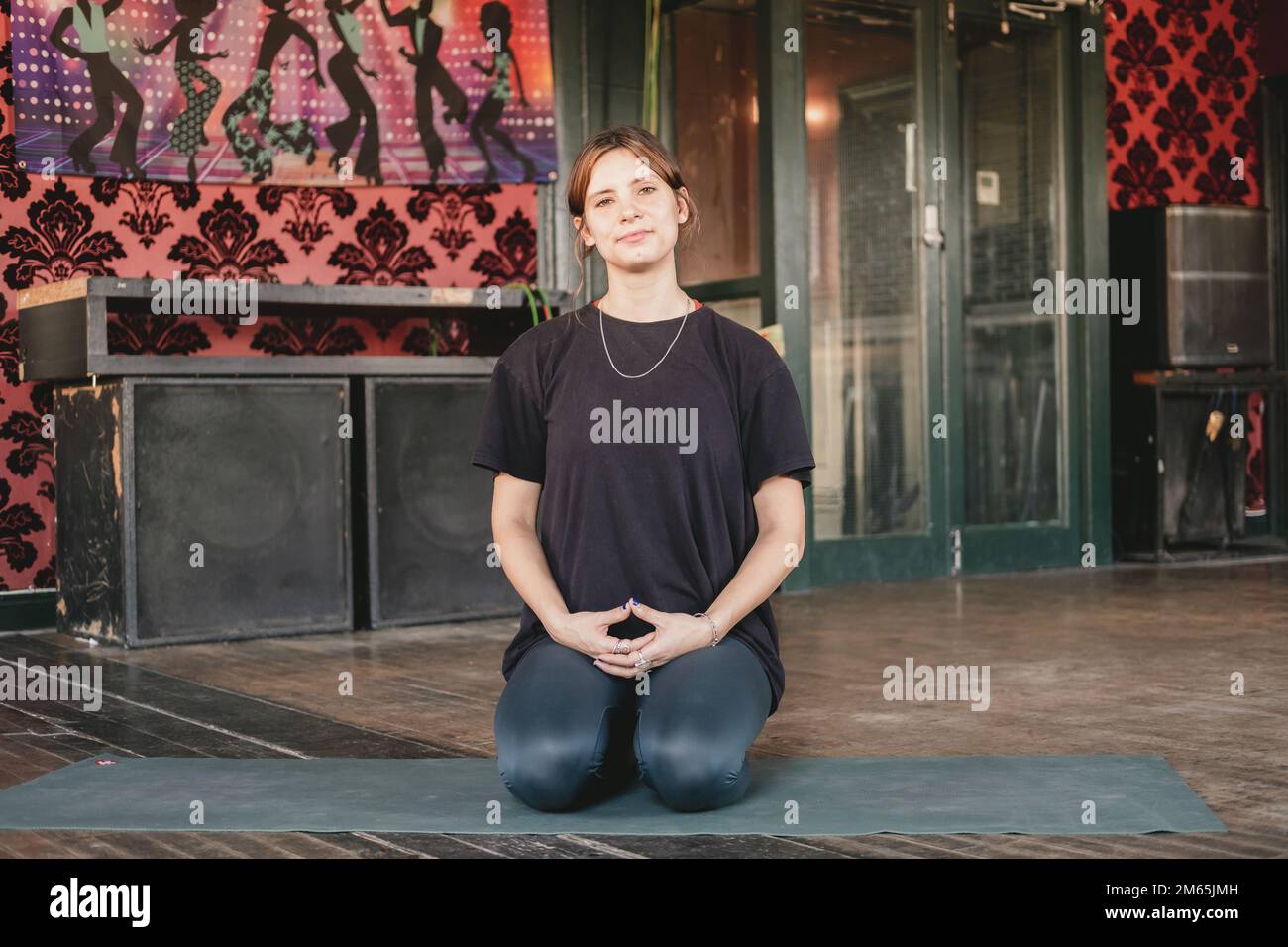 Ganzkörper-Horizontalfoto einer neuen Yoga-Lehrerin, die sich in Kniehaltung ruht (vajrasana) und während ihres Vinyasa-Flow-Yoga-Trainings in die Kamera schaut Stockfoto