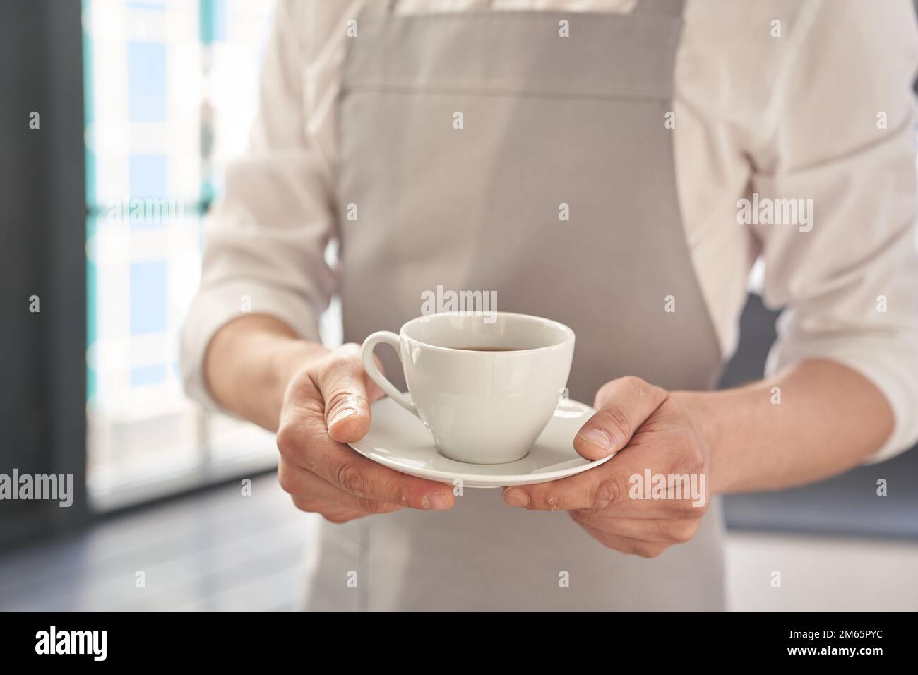 Die Hände des Kellners in einer grauen Schürze halten eine Tasse Kaffee. Der Barista gibt eine Tasse heißen Kaffee in einem Café vor dem Hintergrund eines großen Fensters. Hochwertiges Foto Stockfoto