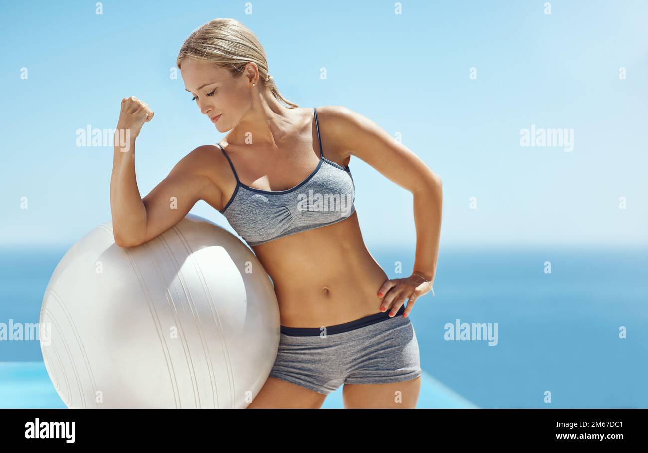 Flexibel. Eine attraktive junge Frau, die nach ihrem Workout ihren Bizeps auf einem Sportball am Pool bewegte. Stockfoto