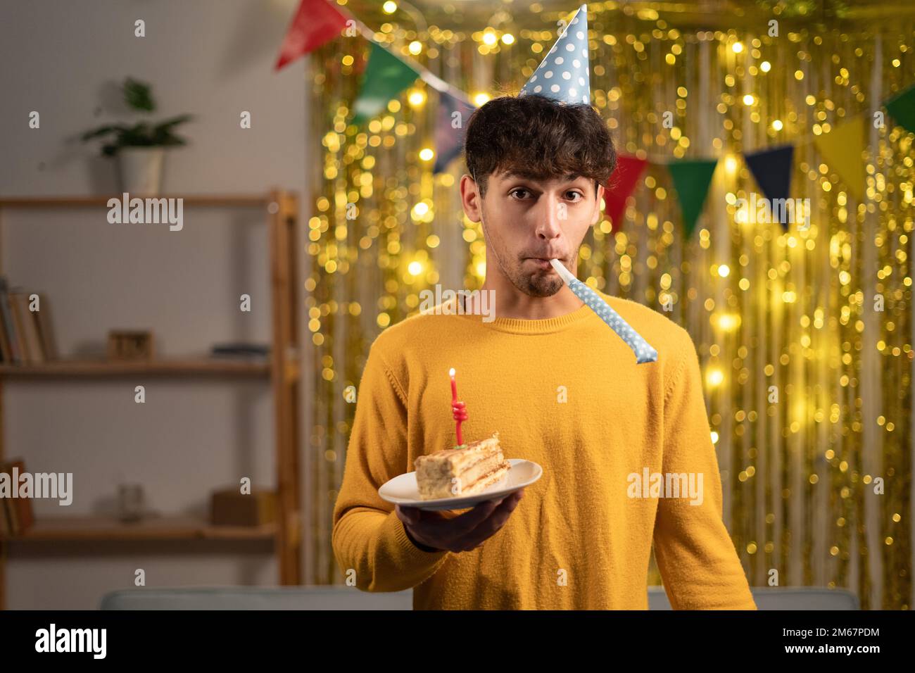 Ein komischer Kerl feiert zu Hause Geburtstag, hat Geburtstagskuchen in der Hand und trägt einen Partyhut, steht über goldenem Folienhintergrund Stockfoto