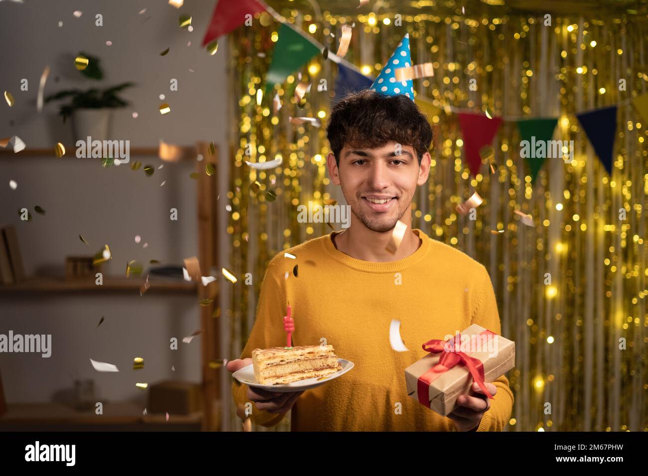 Bild eines gutaussehenden, glücklichen jungen Mannes, der zu Hause seinen Jahrestag feiert und mit Kuchen und Geschenkbox grinselt. Leute, Spaß und Stockfoto