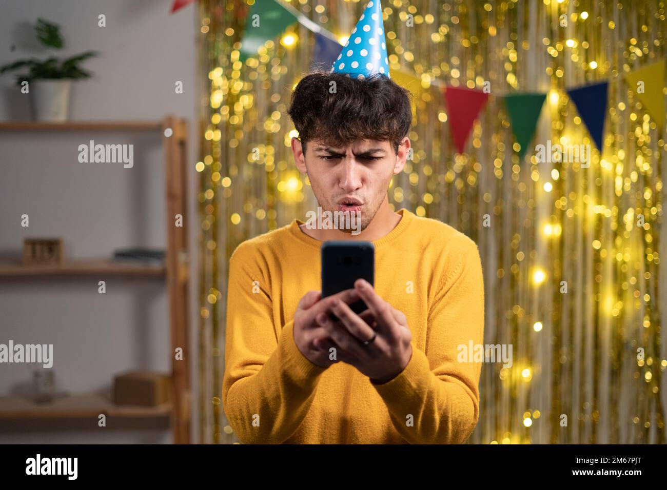 Porträt eines araber mit festlichem Hut, der Handys benutzt, um falsche schlechte Nachrichten zu lesen und seine Geburtstagsparty zu Hause zu feiern Stockfoto