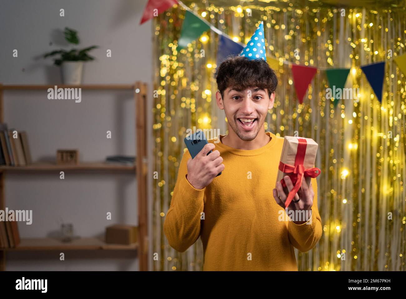 Portrait eines arabers mit festlichem Hut überglücklich fröhlich fröhlich freudig aufgeregter junger Mann, der mit dem Handy tolle Nachrichten liest und eine Geschenkbox hat. Geburtstag zu Hause Stockfoto