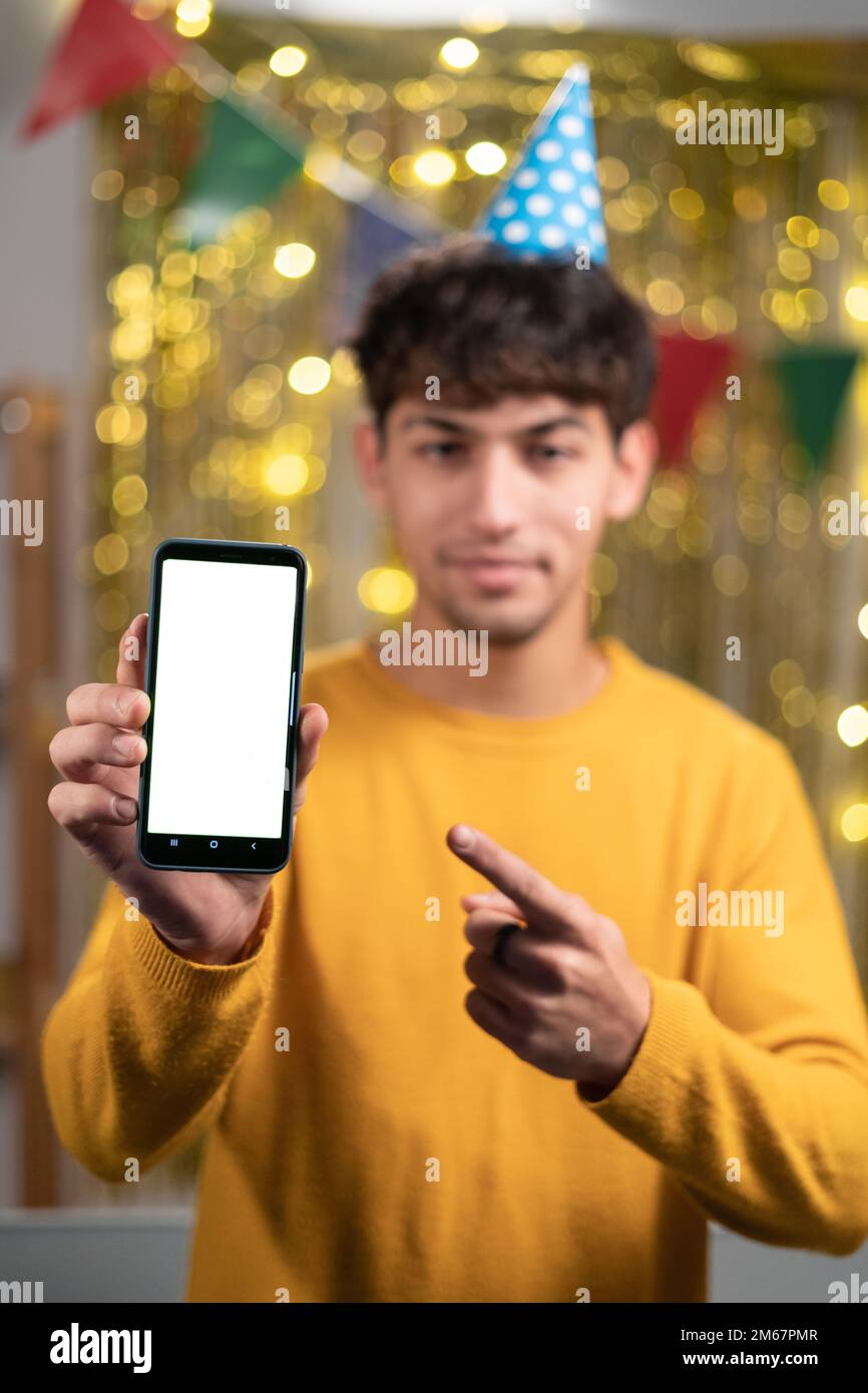 Modellkonzept. Ein gutaussehender, selbstbewusster araber in einem gelben Pullover und einem festlichen Hut, der ein Smartphone mit leerem Bildschirm zeigt und mit dem Finger auf ein leeres Gerät zeigt Stockfoto