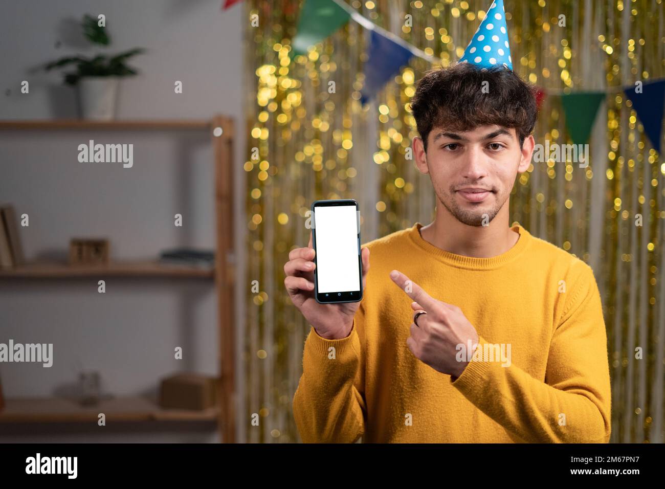 Modell eines jungen Mannes mit festlichem Hut, der mit dem Finger auf ein Mobiltelefon zeigt, mit einem leeren weißen Bildschirm, feiern die Geburtstagsparty zu Hause Stockfoto