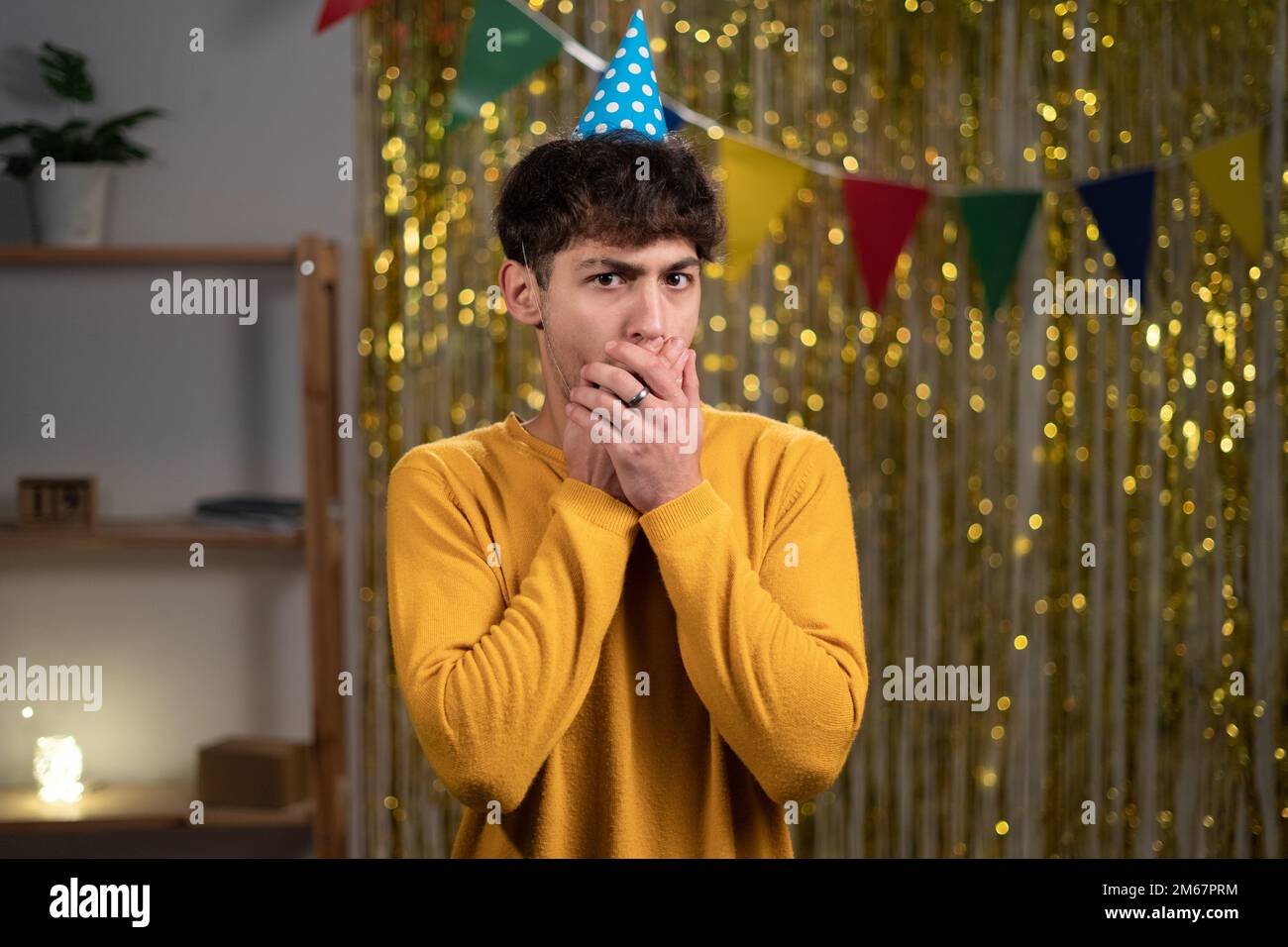 Ein junger, gutaussehender Mann feiert seine Geburtstagsparty zu Hause, schockiert, indem er den Mund mit Händen verdeckt. Geheim Stockfoto