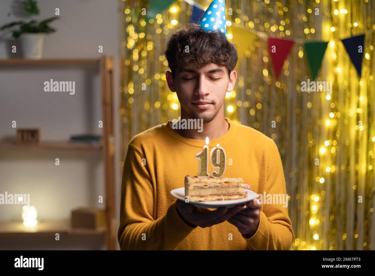 Ein junger Mann mit einem Konus-Partyhut, der sich seinen Geburtstagskuchen mit Kerzen Nummer 19 während der Feier zu Hause wünscht. 19. Geburtstag Stockfoto