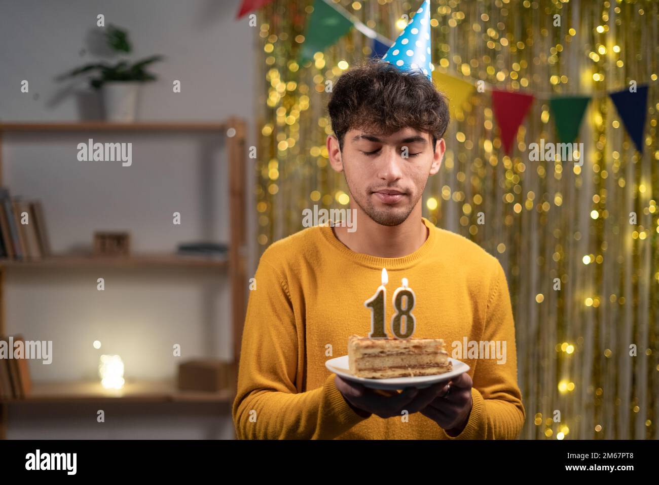 Ein junger Mann mit einem Konus-Partyhut, der sich seinen Geburtstagskuchen mit Kerzen Nummer 18 während der Feier zu Hause wünscht. Geburtstag der Achtzehnten Stockfoto