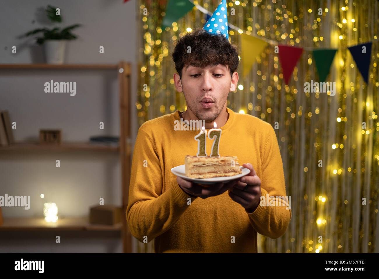 Arabische Schülerin in Cone-Party-Hut wünscht sich und bläst Kerzen Nummer 17 auf Geburtstagskuchen während der Feier zu Hause. Seventeens Geburtstag Stockfoto
