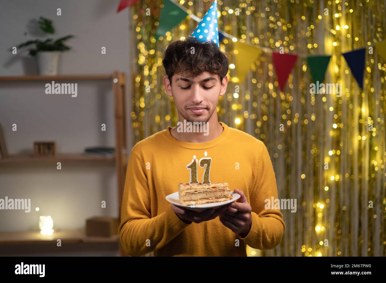 Ein junger Mann mit einem Konus-Partyhut, der sich seinen Geburtstagskuchen mit Kerzen Nummer 17 während der Feier zu Hause wünscht. Seventeens Geburtstag Stockfoto