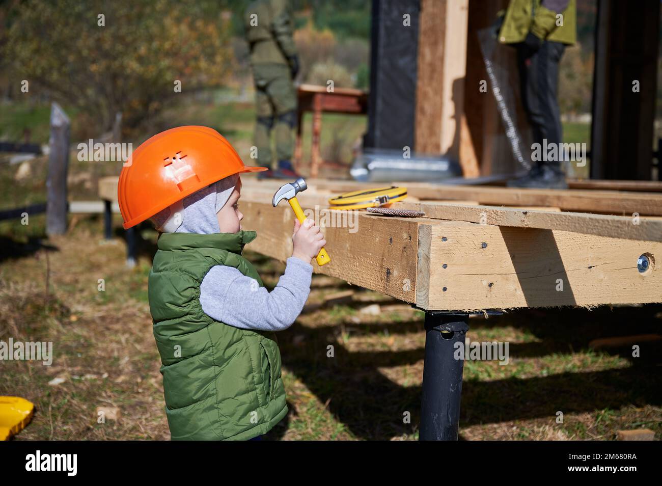 Junge Kleinkinder spielen als Baumeister auf der Baustelle. Zimmermann in orangefarbenem Helm lernt, an sonnigen Tagen ein Haus mit Holzrahmen im Freien zu bauen. Zimmerei- und Werkstattkonzept. Stockfoto