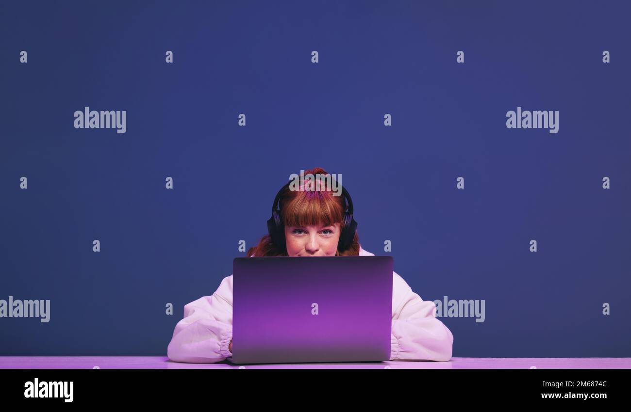 Eine Frau mit Ingwerhaar, die in die Kamera schaut, während sie in einem Studio hinter ihrem Gaming-Laptop sitzt. Weibliche Gamer, die einen interaktiven Streaming-Dienst verwenden w Stockfoto