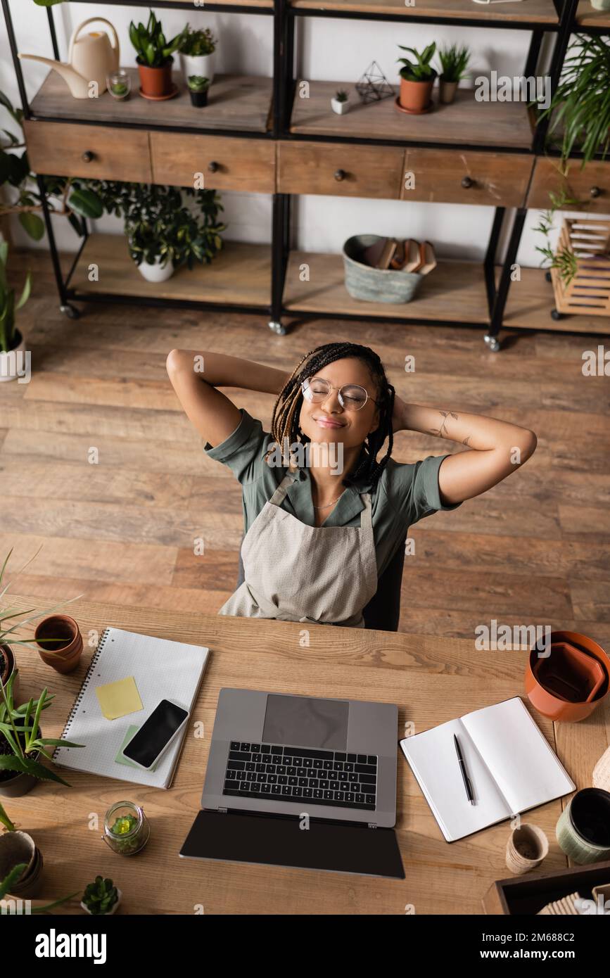 Blick von oben auf den frohen afroamerikanischen Floristen, der mit geschlossenen Augen und Händen hinter dem Kopf neben Geräten und Topfpflanzen im Geschäft sitzt, Bild von Stock Stockfoto