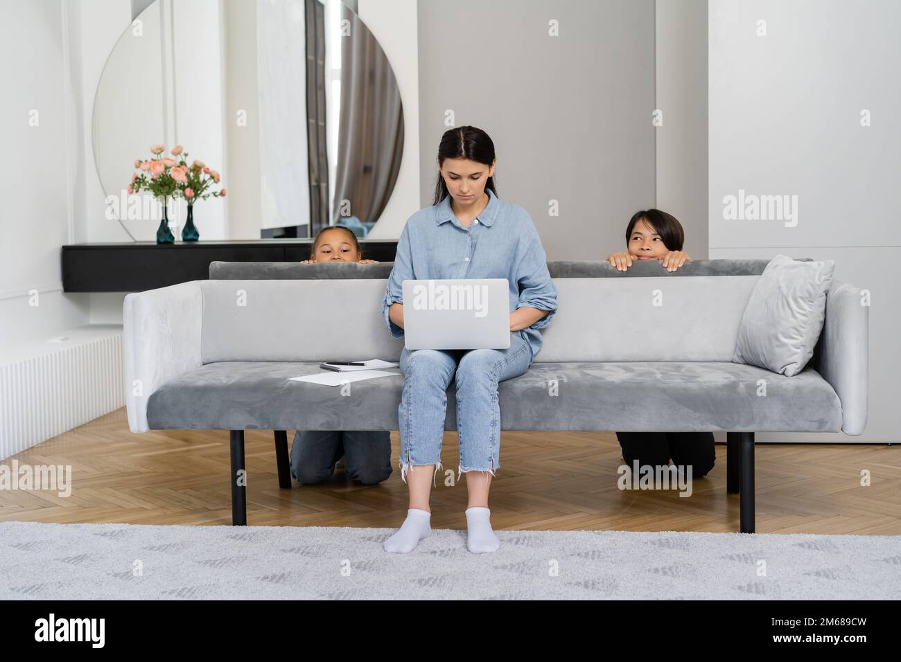 Frau, die zu Hause an einem Laptop arbeitet, fast schlaue asiatische Kinder hinter der Couch, Stockbild Stockfoto