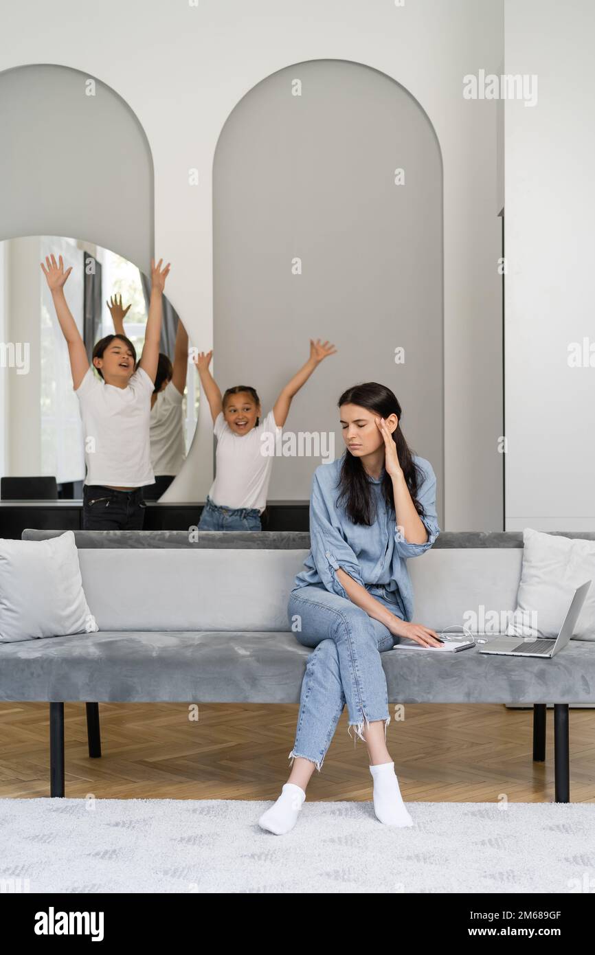 Müde Frau sitzt neben dem Laptop und fröhliche asiatische Kinder zu Hause, Stock Image Stockfoto