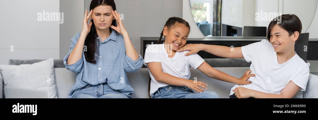 Frau mit Kopfschmerzen in der Nähe von fröhlichen asiatischen Kindern zu Hause, Banner, Stock Image Stockfoto