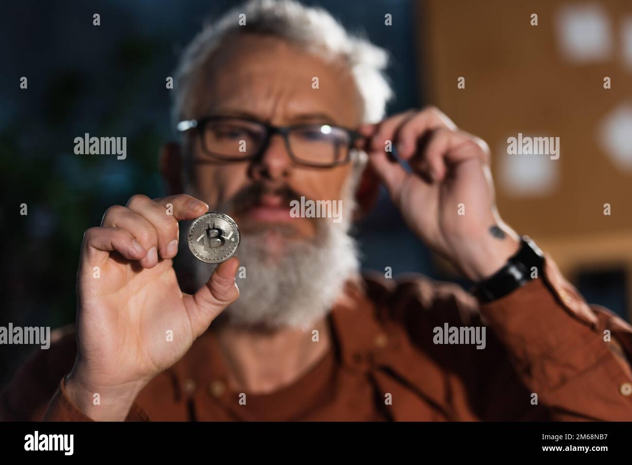 KIEW, UKRAINE - 17. NOVEMBER 2022: Unscharfer, bärtiger Geschäftsmann, der die Brille anpasst, während er Silberbitmünze in der Hand hält Stockfoto