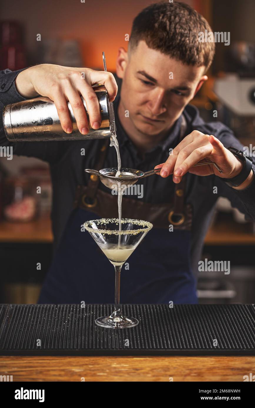 Der Barkeeper filtert den alkoholischen Cocktail vorsichtig aus dem Schüttelbecher aus Stahl durch das Sieb in das Glas. Stockfoto