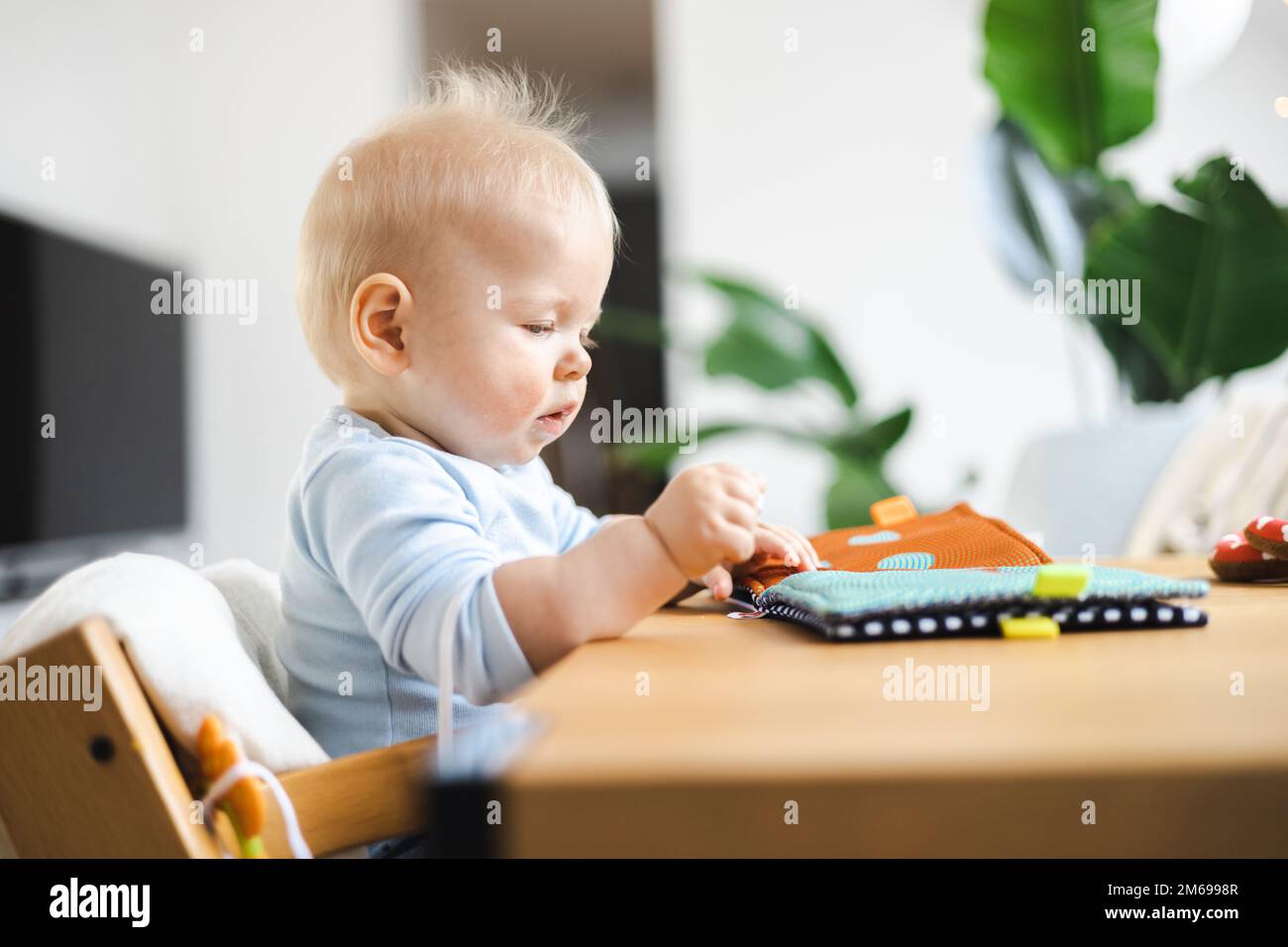 Glückliches Kind sitzt am Esstisch und spielt mit seinem Spielzeug in einem traditionellen skandinavischen Designer-Hochstuhl aus Holz in einem modernen, hellen Wohnhaus. Süßes Baby, das mit Spielzeug spielt. Stockfoto