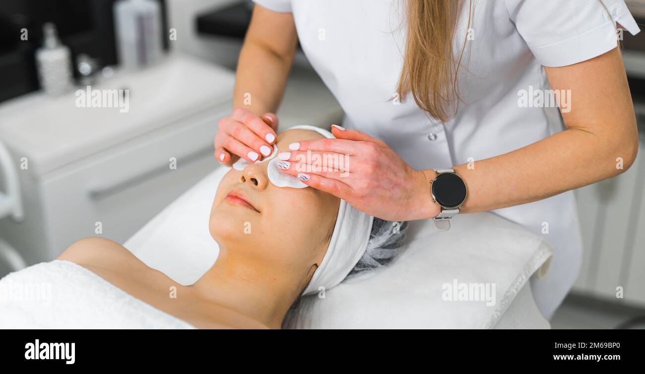 Eine Frau in einem Handtuch, die sich hinlegt, während eine Kosmetikerin ihr eine Augenkompression ins Gesicht legt. Schönheitskonzept. Hochwertiges Foto Stockfoto