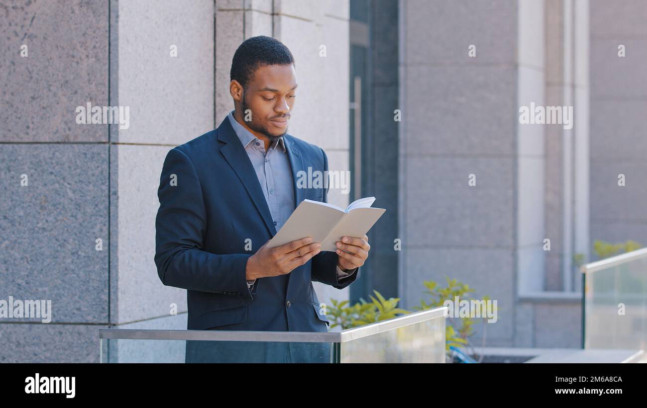 Afroamerikanischer Mann, ethnischer Geschäftsmann, Arbeitgeber, Unternehmer, Büroangestellter, der auf dem Balkon steht, mit einem Tagesplaner, der Papier liest, Bücher liest, Geschäfte liest Stockfoto