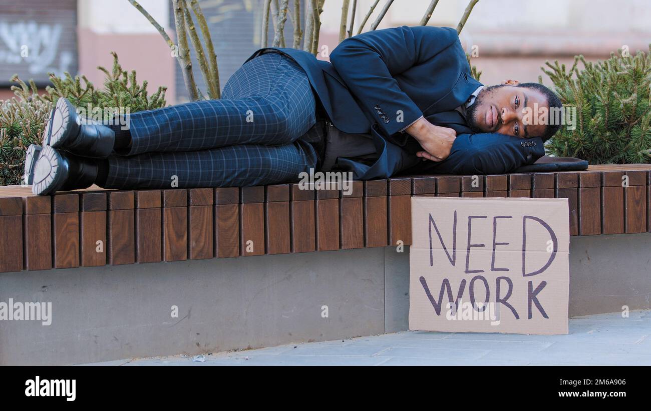 Schlafende Obdachlose gefeuerte afroamerikanische Männer schlafen auf einer Bank in der Stadt mit Poster brauchen Arbeit. Ein einsamer, aufgebrachter, ethnischer Typ verlor im Park seinen Job Stockfoto