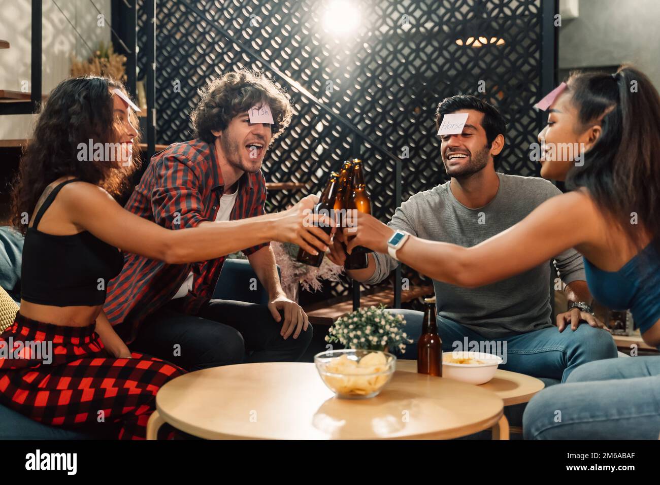 Eine Gruppe glücklicher männlicher und weiblicher Freunde, die Namensspiel spielen und mit einer Bierflasche anstoßen, während sie zu Hause während der Hausarming-Party sitzen Stockfoto