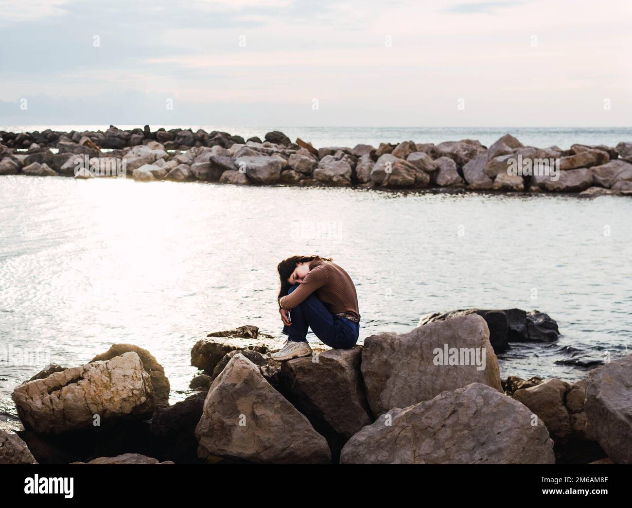 Eine Frau, die auf Steinen vor dem Meer sitzt und bei Sonnenuntergang ihre Beine umarmt Stockfoto