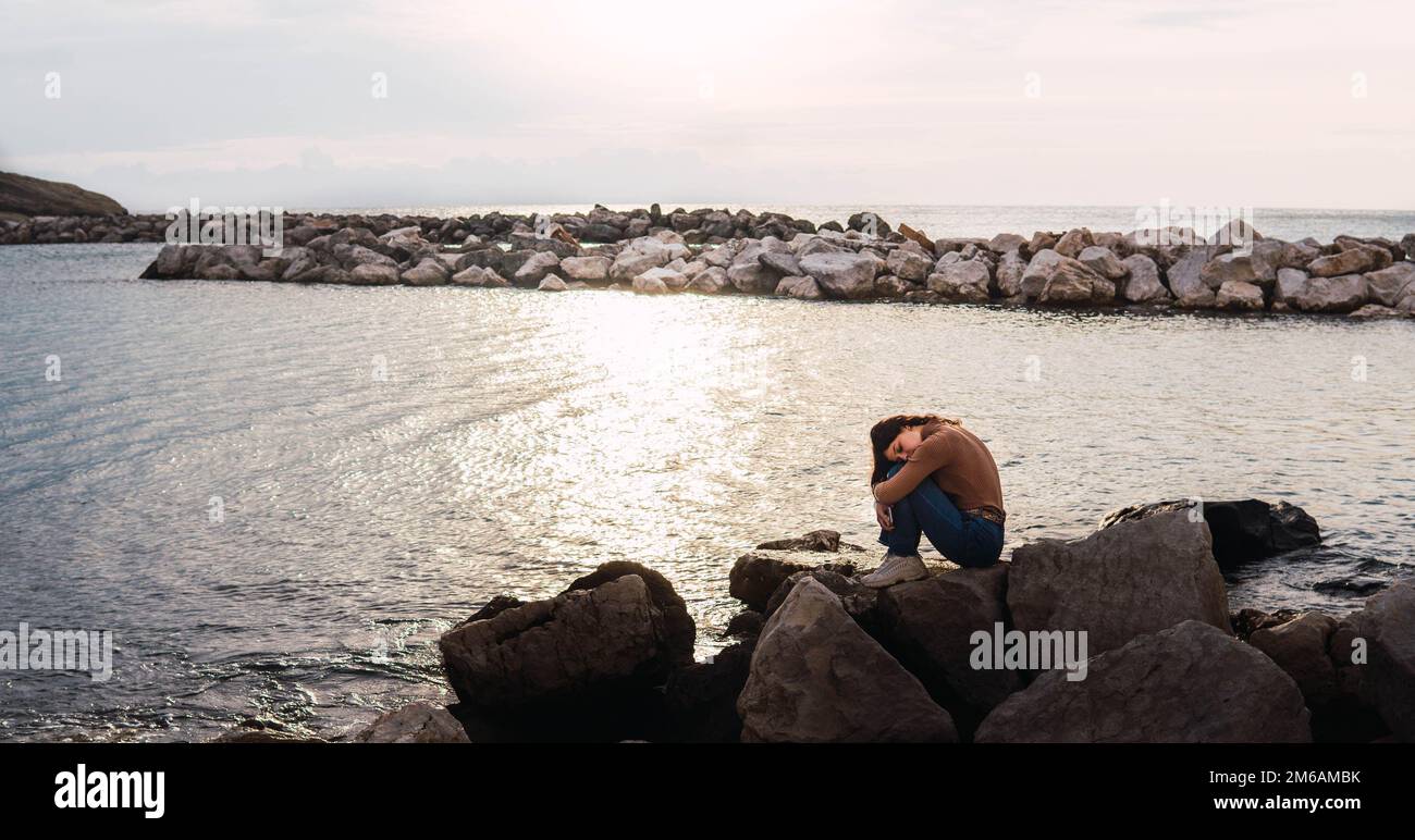 Eine Frau, die auf Steinen vor dem Meer sitzt und bei Sonnenuntergang ihre Beine umarmt Stockfoto