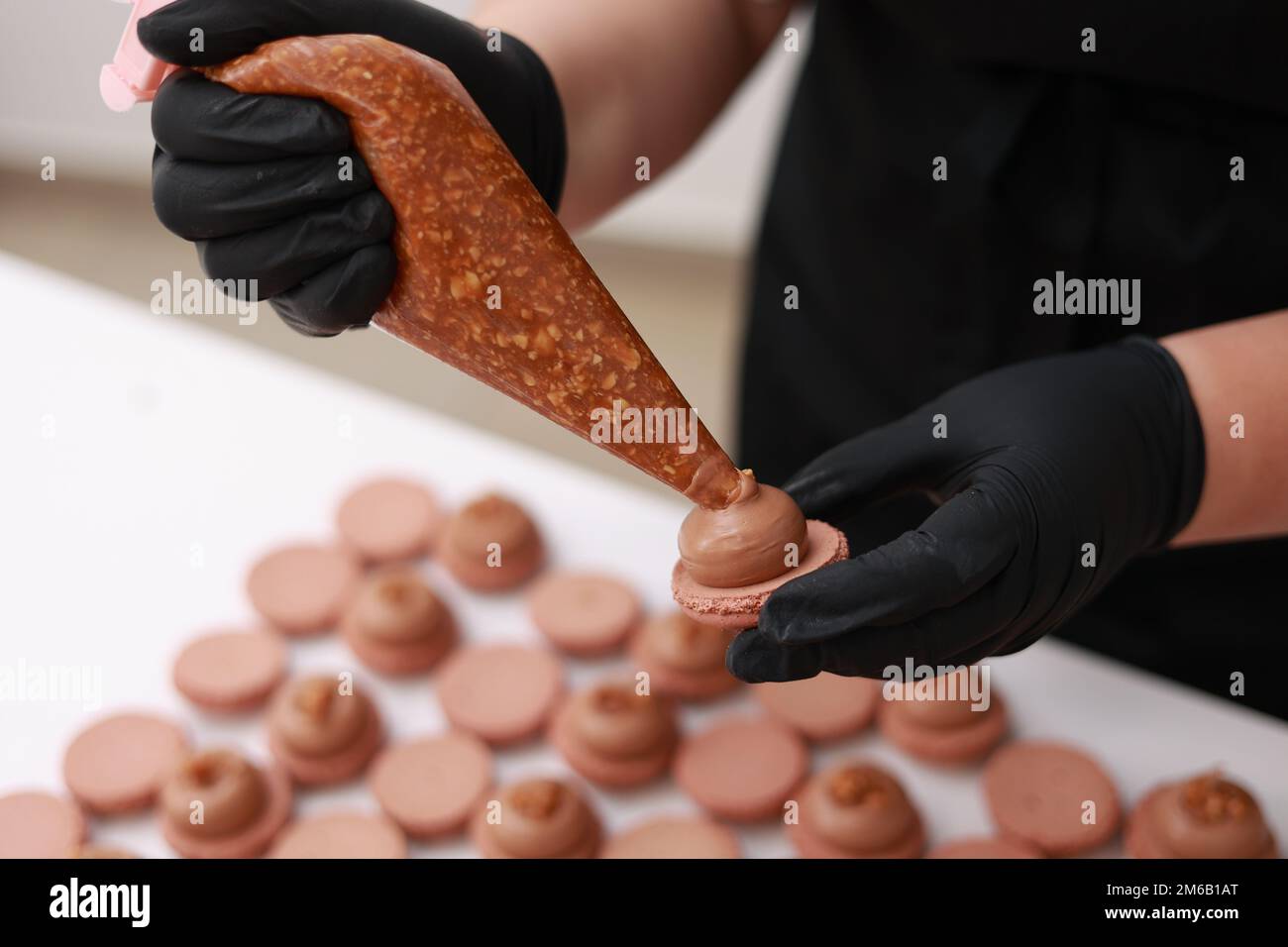 Nahaufnahme von Händen des Küchenchefs, die Salz-Karamell mit Nüssen in Ganache-Creme auf Schokoladenmakronen-Muscheln schütten. Makronen-Backen-Konzept. Kochen, Essen Und Backen Stockfoto