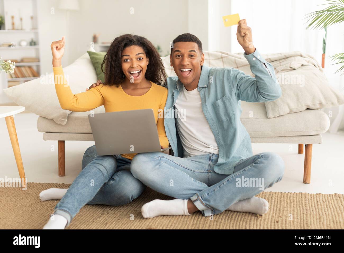 Lachender afroamerikanischer Millennials umarmt eine Frau, benutzt Laptop, Kreditkarte, hebt die Hand, macht Sieg und Erfolg Geste Stockfoto