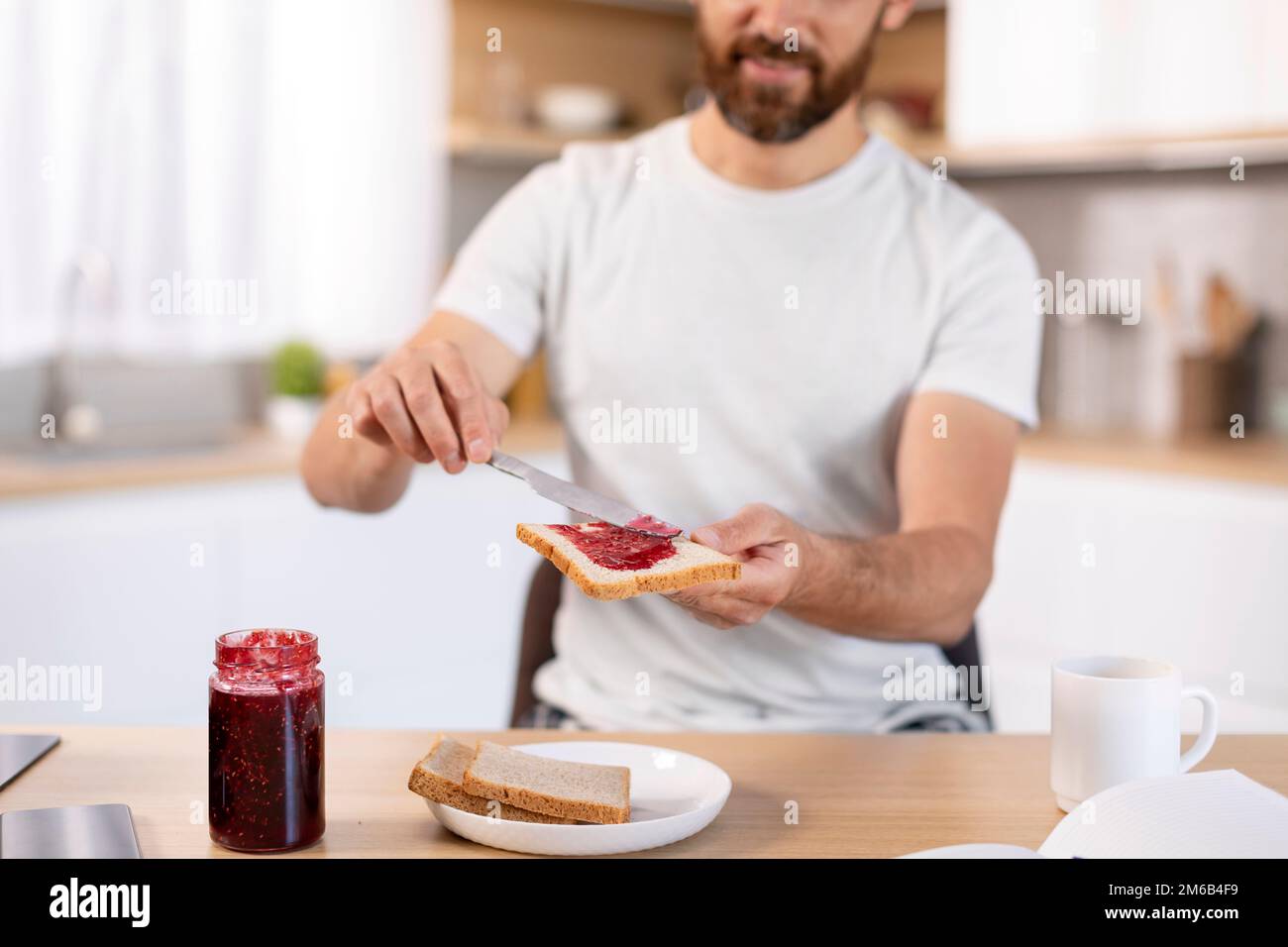 Lächelnder erwachsener Kaukasier mit Bart, der Sandwich mit Marmelade am Tisch macht, genießt das Wochenende in der Küche Stockfoto