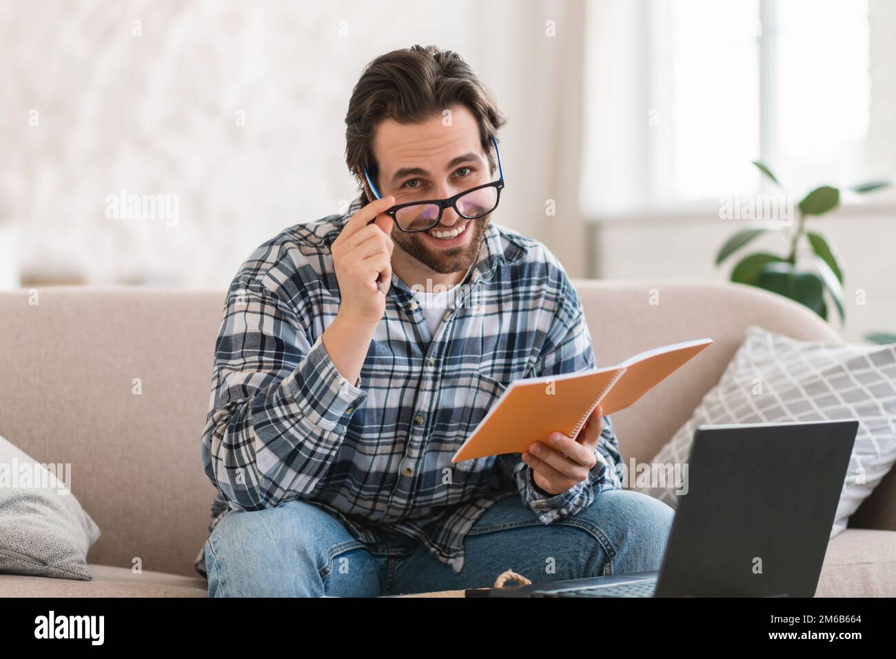 Lustig lächelnder junger weißer Lehrer nimmt die Brille ab, schaut in die Kamera, sitzt auf dem Sofa mit Computer Stockfoto