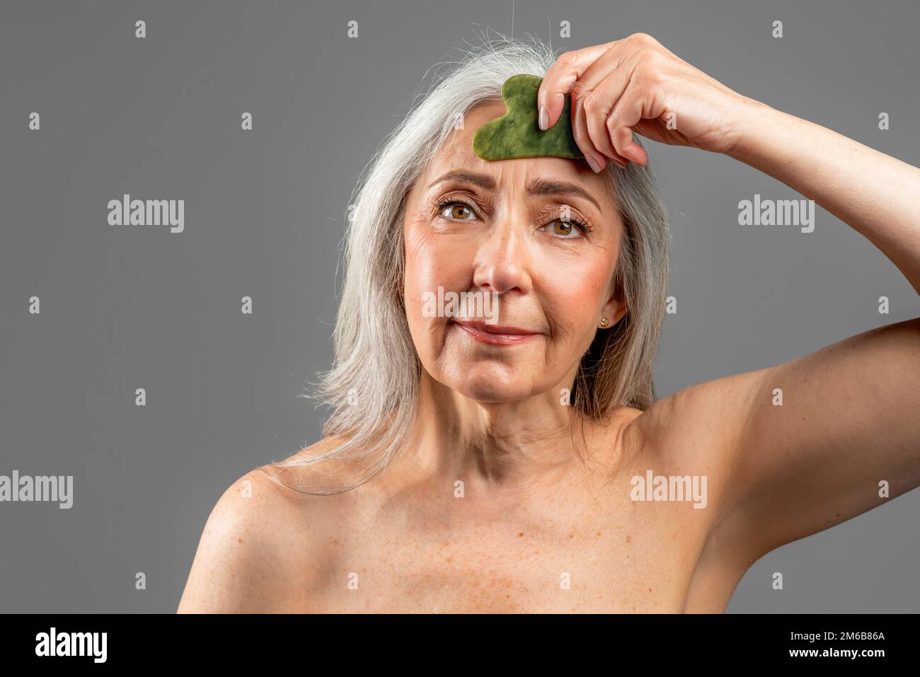 Ernsthaft pensionierte europäische Frau mit grauen Haaren, die Jade-Massage mit Gua-Sha-Stein macht, isoliert auf grauem Hintergrund Stockfoto