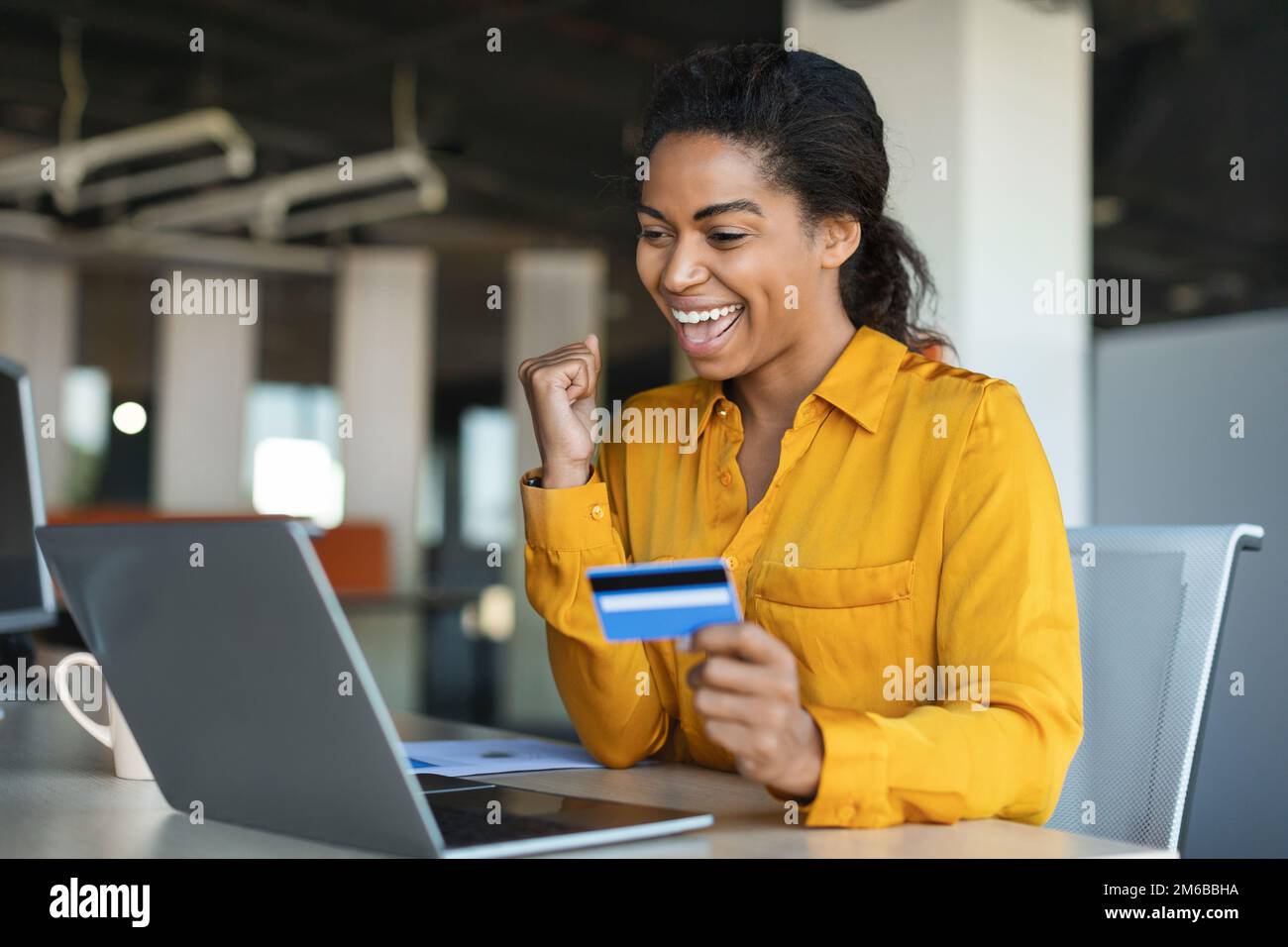 Eine überfreuliche afroamerikanische Geschäftsfrau, die eine erfolgreiche Geste macht, eine Kreditkarte in der Hand hält, mit einem Laptop am Tisch sitzt Stockfoto