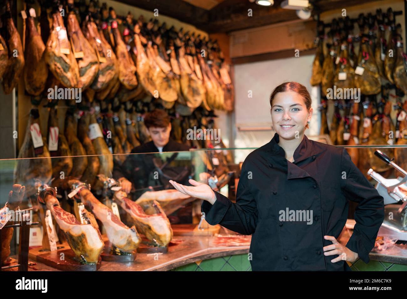 Lächelnde Verkäuferin in schwarzer Uniform, die iberischen Schinken im Fleischladen empfiehlt Stockfoto