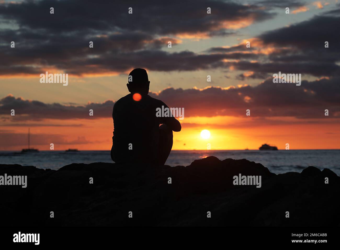 Ein Mann, der allein auf Felsen am Strand sitzt und den Sonnenuntergang anschaut, in Gedanken verloren, eine Überlegung Stockfoto