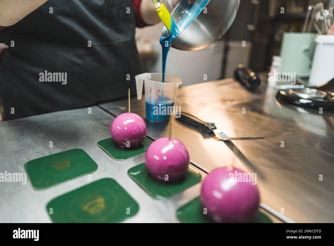 Ein professioneller Bäcker mit schwarzer Schürze, der blaue Glasur in eine Tasse gießt, bildet eine Metallschüssel. Drei Kuchen mit pinkfarbenem Glasur neben dem Becher. Verschwommener Hintergrund. Hochwertiges Foto Stockfoto
