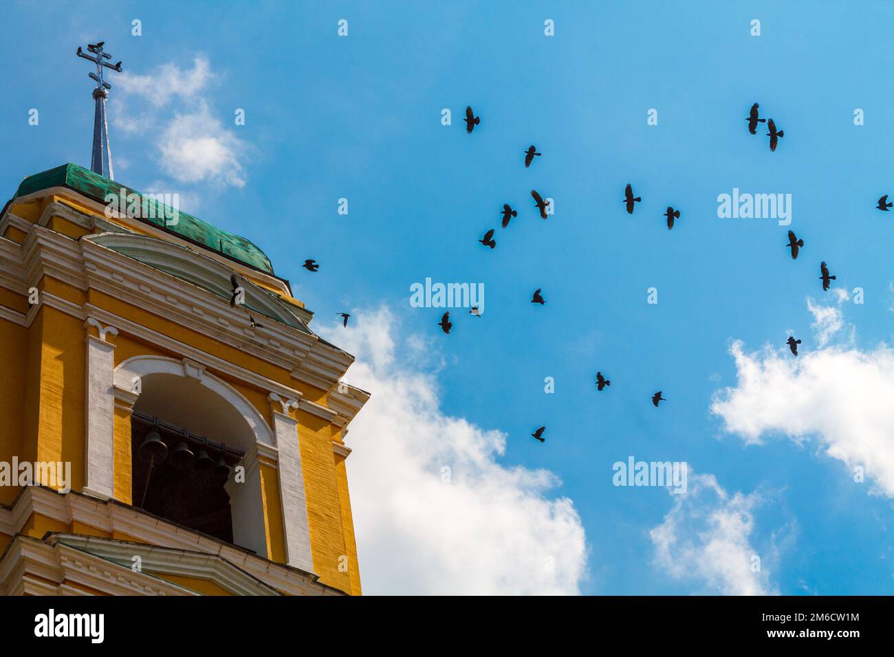 Turm des christlichen Tempels mit Glocken und einem Kreuz, in das viele schwarze Vögel gegen den blauen Himmel mit weißen Wolken fliegen Stockfoto