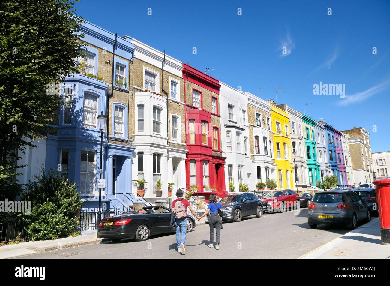 Ein junges Paar, das an einem sonnigen Tag Händchen hält und eine Straße mit bunten Reihen von Reihenhäusern überquert, Notting Hill, London, England, Großbritannien Stockfoto