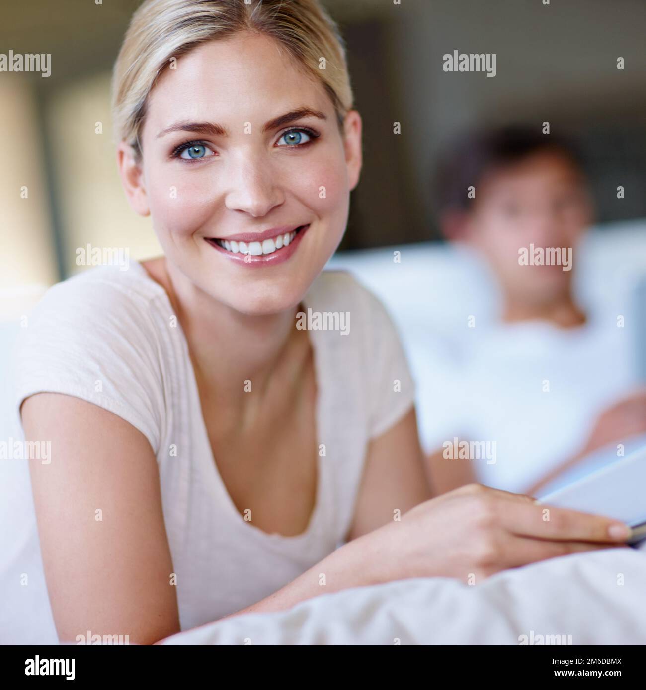 Glück ist ein Insider-Job. Eine glückliche junge Frau lag im Bett und las eine Zeitschrift mit ihrem Mann im Hintergrund. Stockfoto