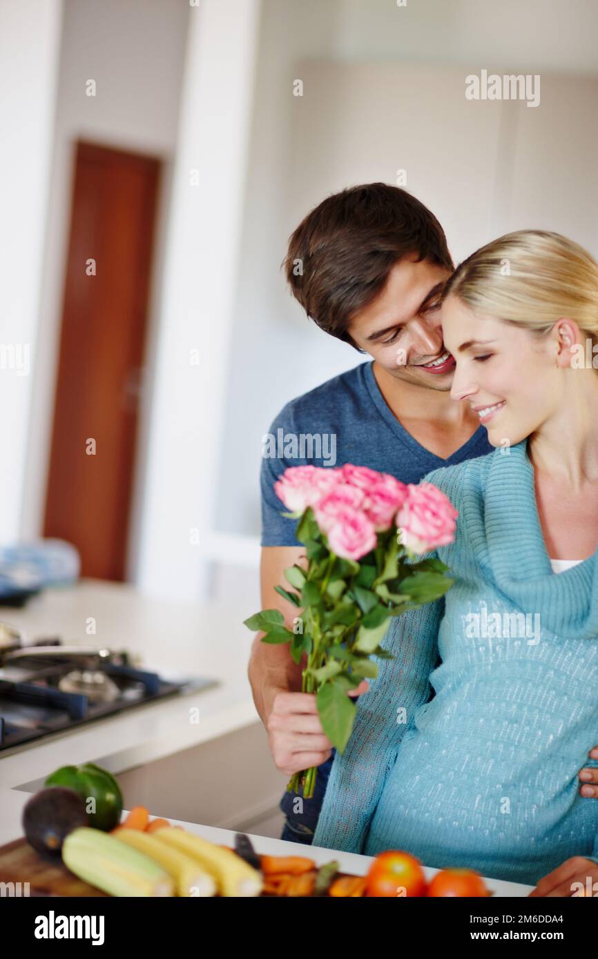 Sie kommen aus meinem Herzen. Ein gutaussehender junger Mann, der seiner Frau einen Strauß rosa Rosen gibt, während sie ein Essen zubereitet. Stockfoto
