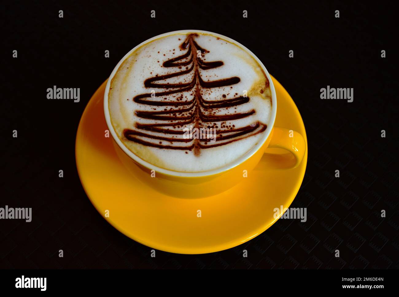 Schaumiger Cappuccino. Isolierter gelber Becher. Dekorative Weihnachtsbaumschmuck auf weißem Schaum. Gourmet-Kaffee. Brauner Schokoladenkiefernbaum. Schwarzer Hintergrund Stockfoto