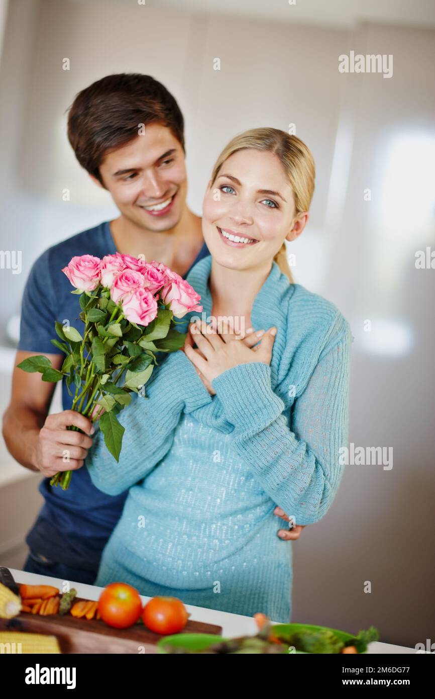 Er ist so gut zu mir. Ein gutaussehender junger Mann, der seiner Frau einen Strauß rosa Rosen gibt, während sie ein Essen zubereitet. Stockfoto